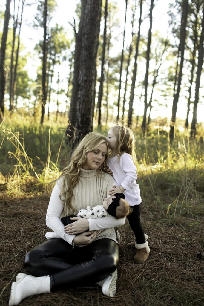 Surrounded by towering pine trees, a mother cradles her newborn while showering her 3-year-old with affection, capturing a beautiful family bond in Decatur