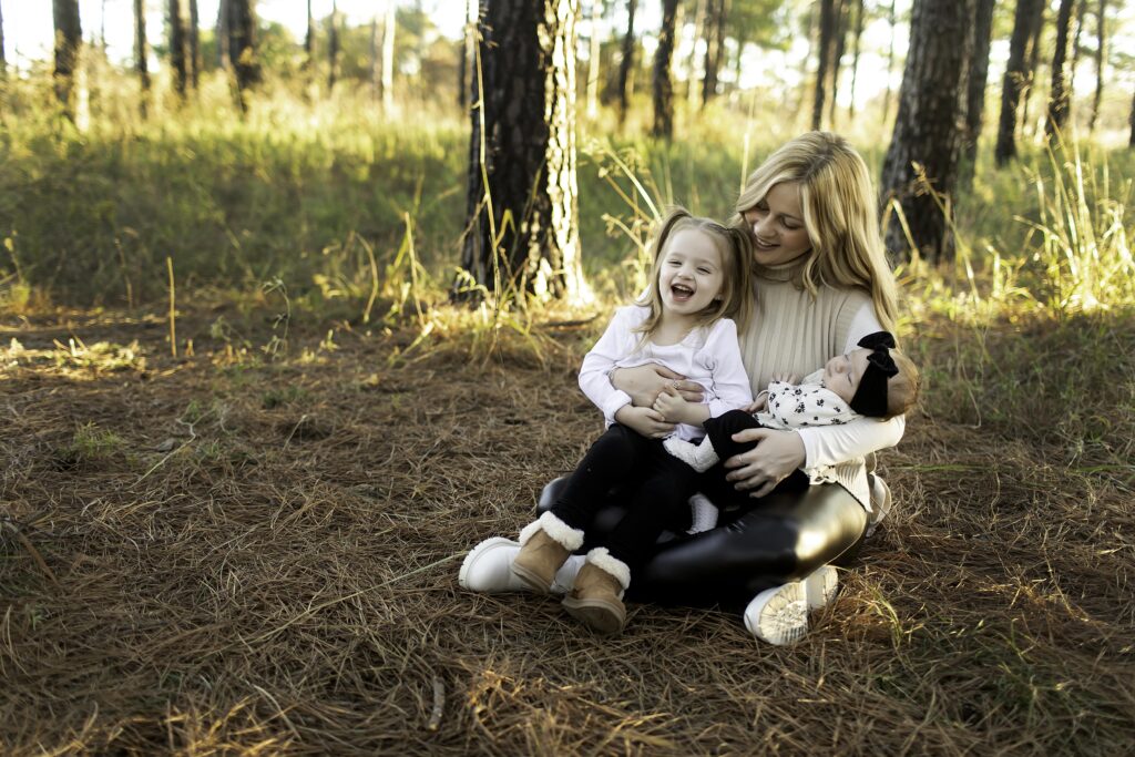 A mom lovingly holds her newborn while her 3-year-old joyfully embraces her, surrounded by the peaceful pine trees of Decatur, creating a moment full of love and connection