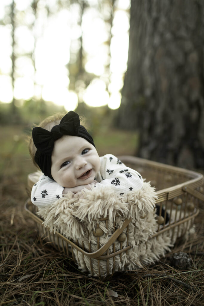 A 2-month-old baby smiles gently while resting in a wicker basket, surrounded by the peaceful pine trees of Decatur, TX, radiating happiness and tranquility