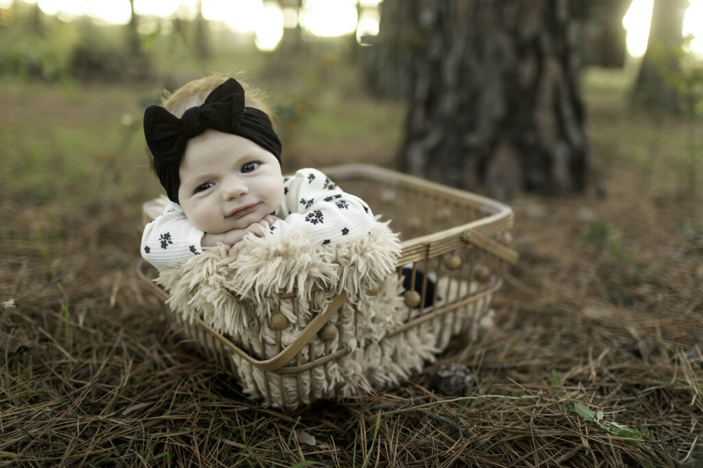 Captured in Decatur’s pine forest, a 2-month-old smiles softly while nestled in a cozy basket, her joy shining through amidst the natural beauty