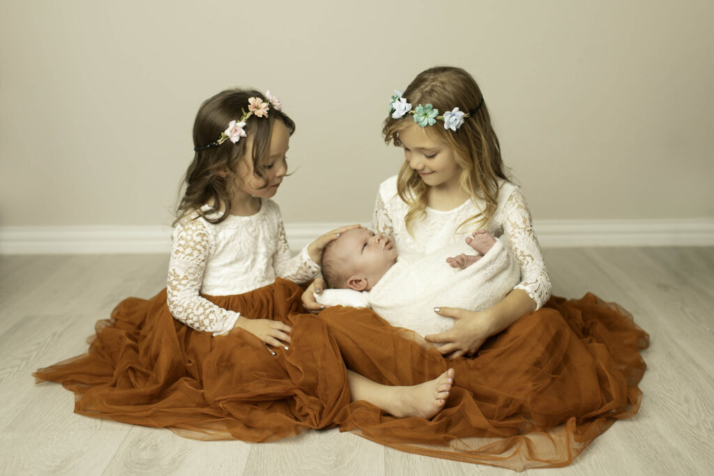 Two sisters gently holding their newborn brother, smiling together in a heartwarming family momen