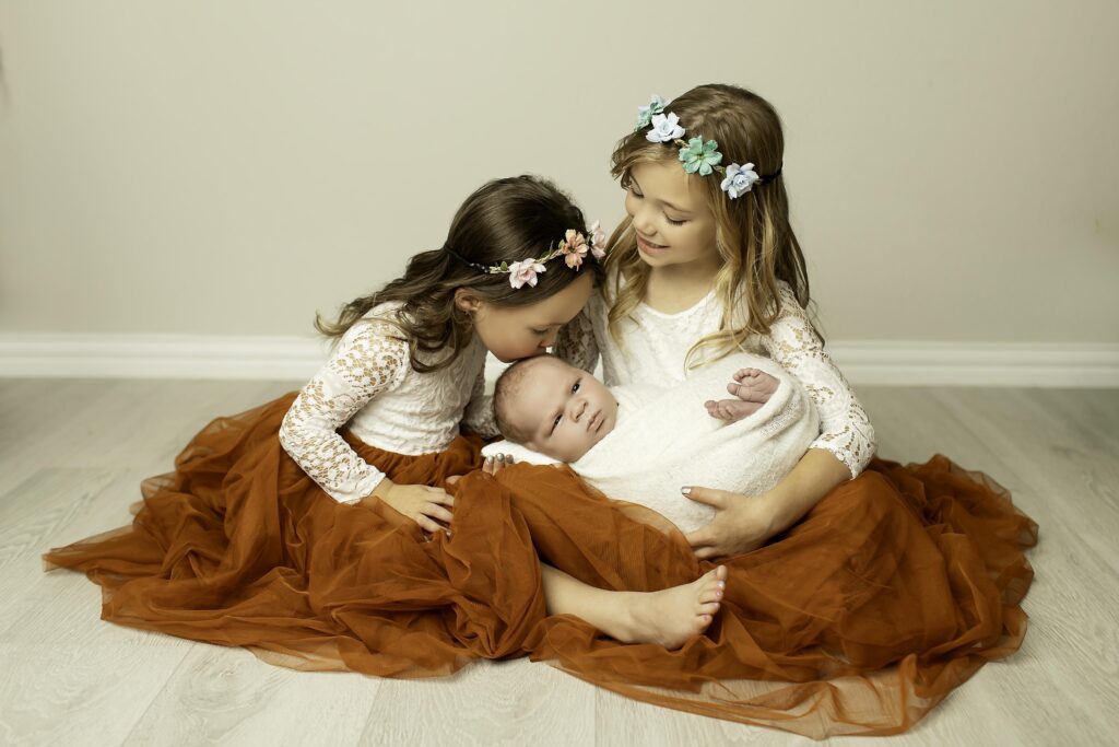 Two sisters gently holding their newborn baby brother, sharing a precious moment together in a cozy setup during a Chunky Monkey session