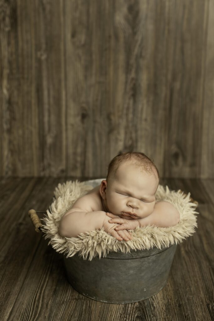 Adorable newborn boy curled up in a vintage bucket, surrounded by soft fabrics and gentle lighting