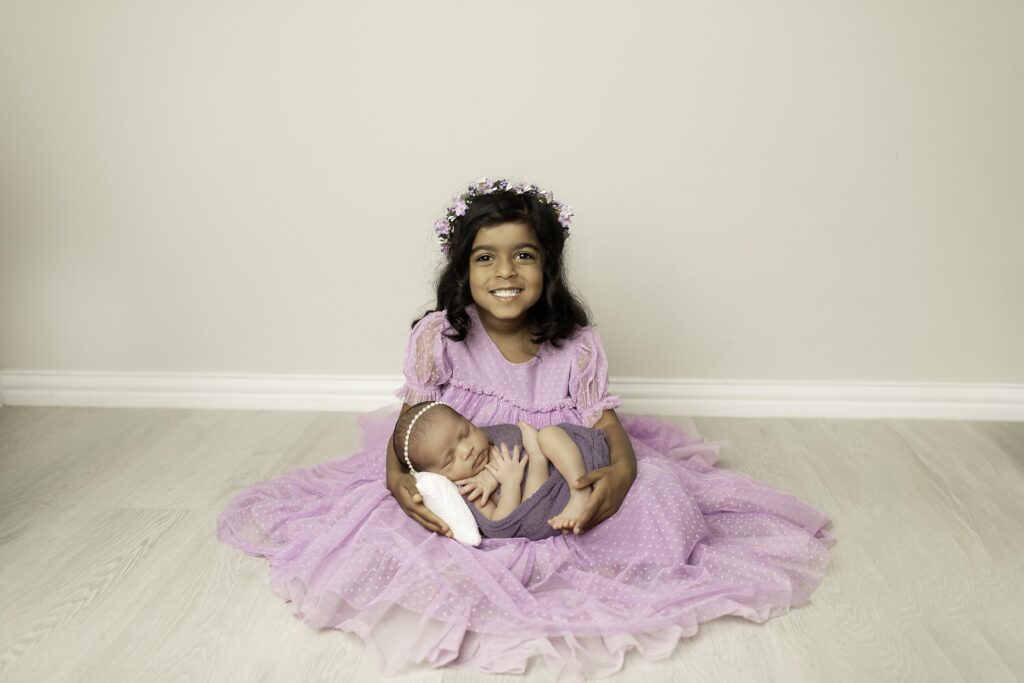 Sweet portrait of a newborn girl with her big sister in a purple dress, sharing a tender moment in the studio