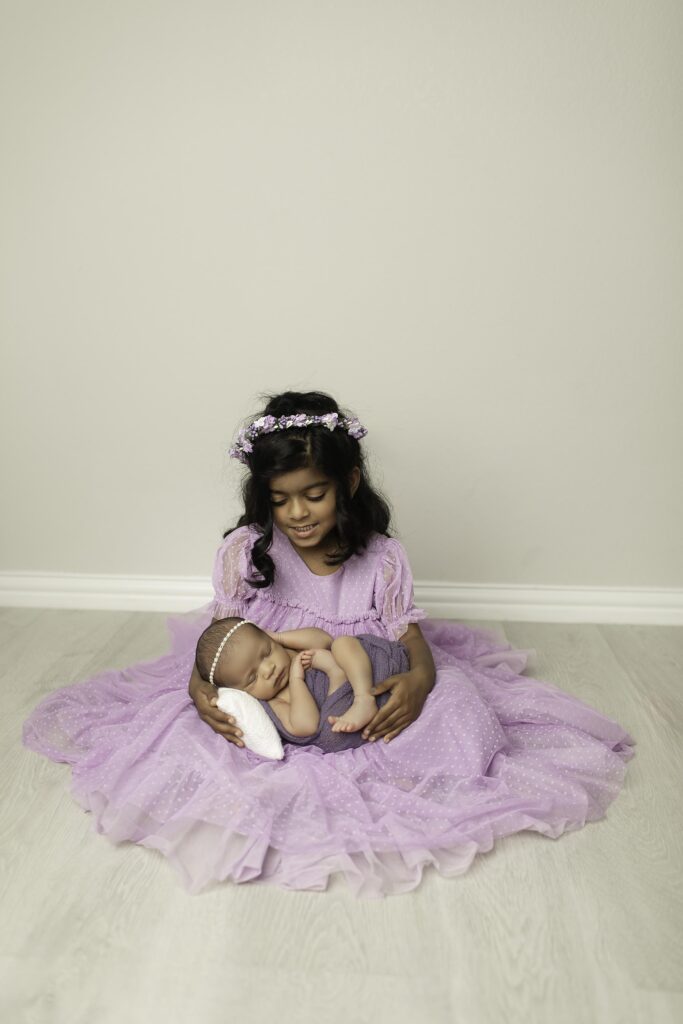 Big sister in a purple dress gently holding her newborn sister, captured in a loving moment in the studio