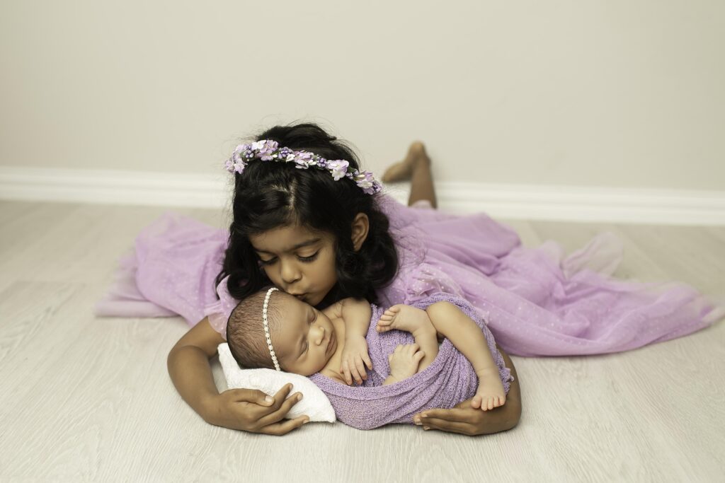 Newborn girl peacefully resting with her big sister, who’s wearing a beautiful purple dress in the studio