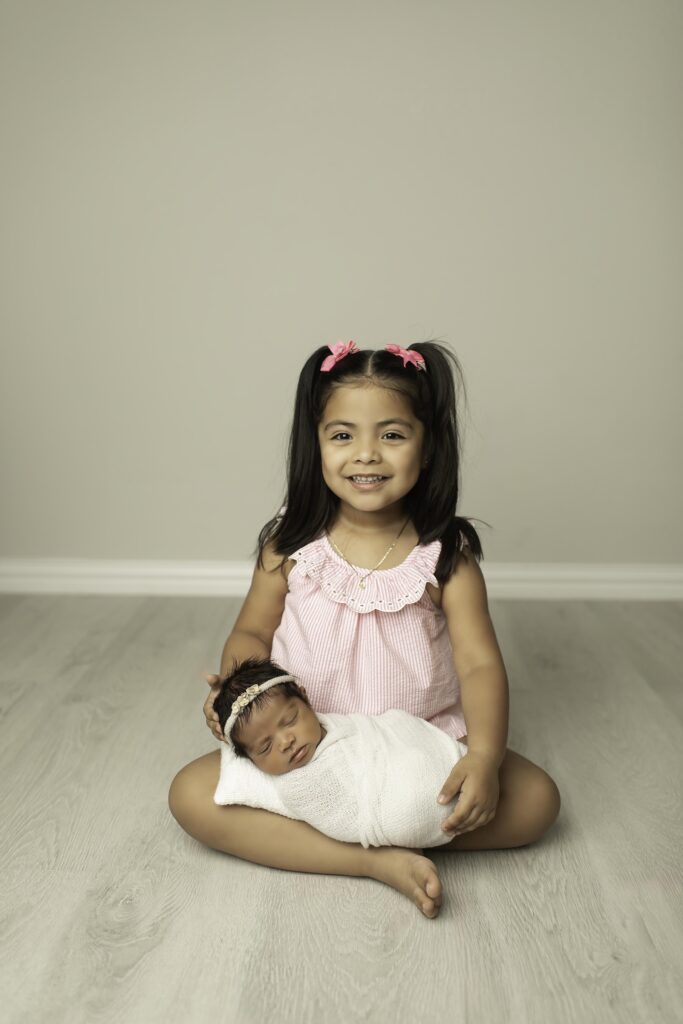 Big sister lovingly holds her newborn sister, both smiling softly as they share a tender moment together in the Fort Worth studio, surrounded by soft lighting