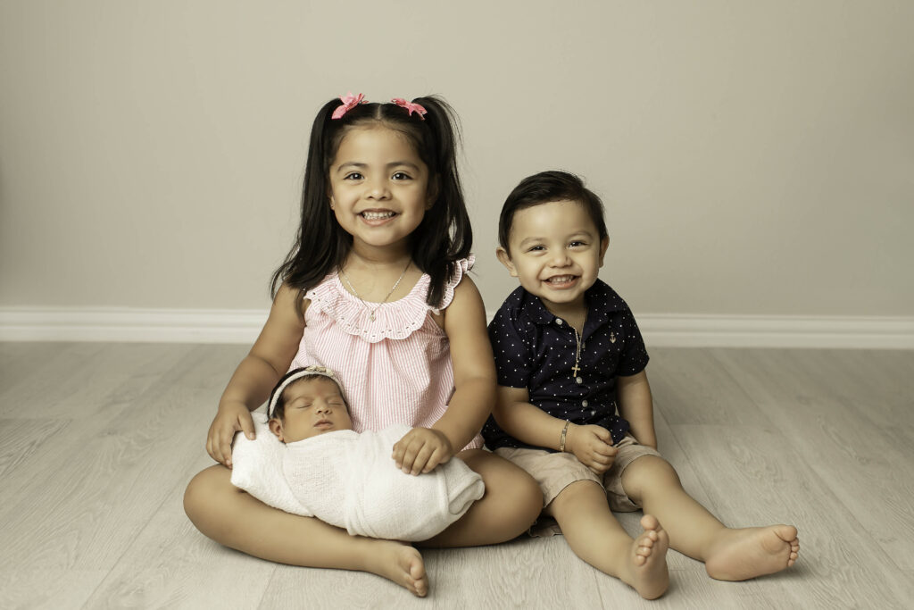 Big sister and big brother gently surround their newborn sibling, all three sharing a joyful, loving moment in the Fort Worth studio, their bond beautifully captured.