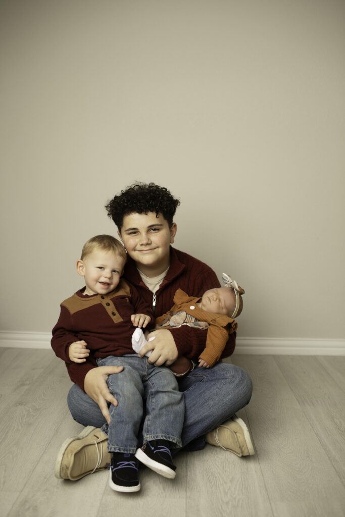 "A heartwarming moment as two big brothers gently hold their newborn sister with chunky cheeks, all three sharing a sweet and tender moment together