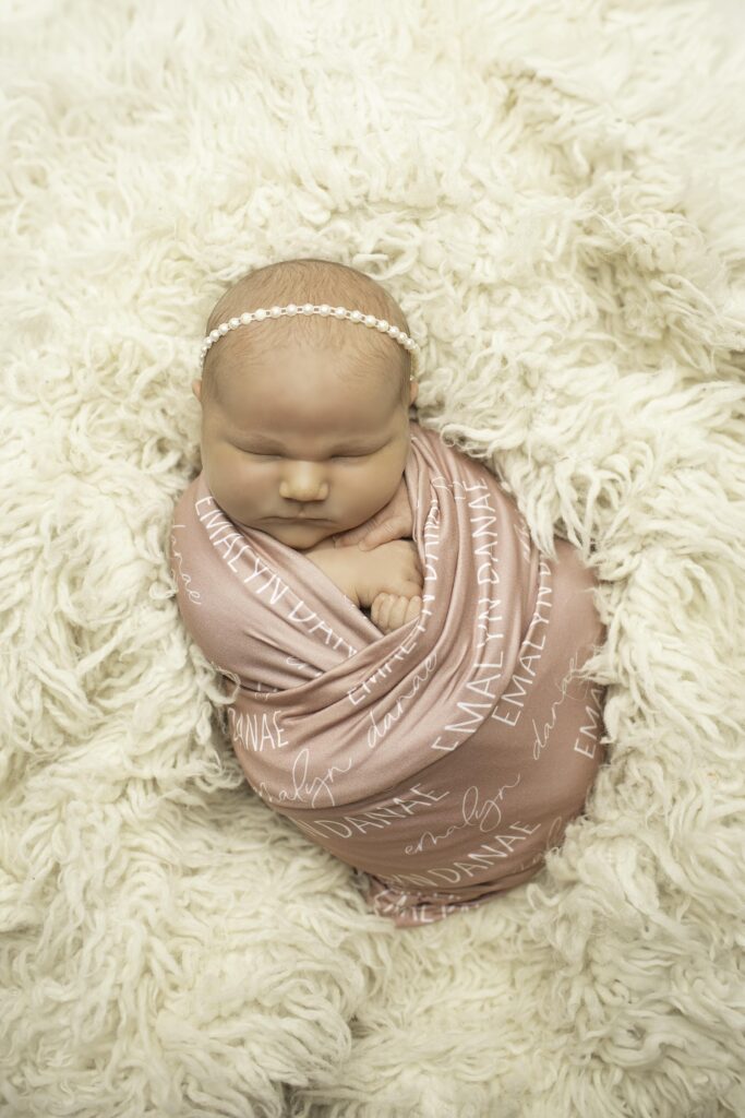 A newborn peacefully sleeps, wrapped snugly in a soft blanket with their name embroidered on it, highlighting their adorable, chunky cheeks