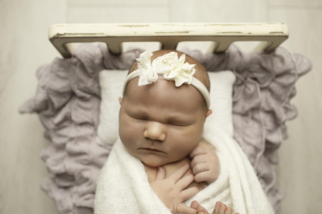 A newborn with soft, chunky cheeks is gently posed on a cozy newborn bed, wrapped in a delicate blanket, peacefully resting during their photoshoot