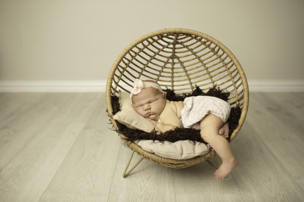 Posed in a wicker basket on their tummy, a newborn with adorable, round cheeks sleeps soundly, surrounded by soft textures and cozy fabrics