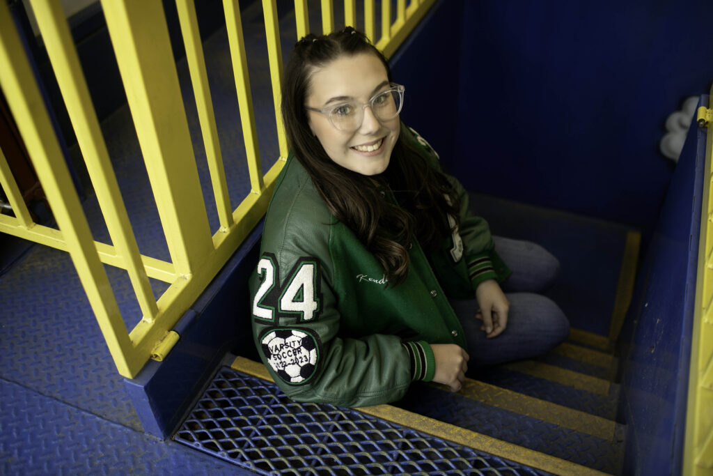 Senior girl posing inside a walk-through funhouse, with playful lighting and bold patterns reflecting around her.