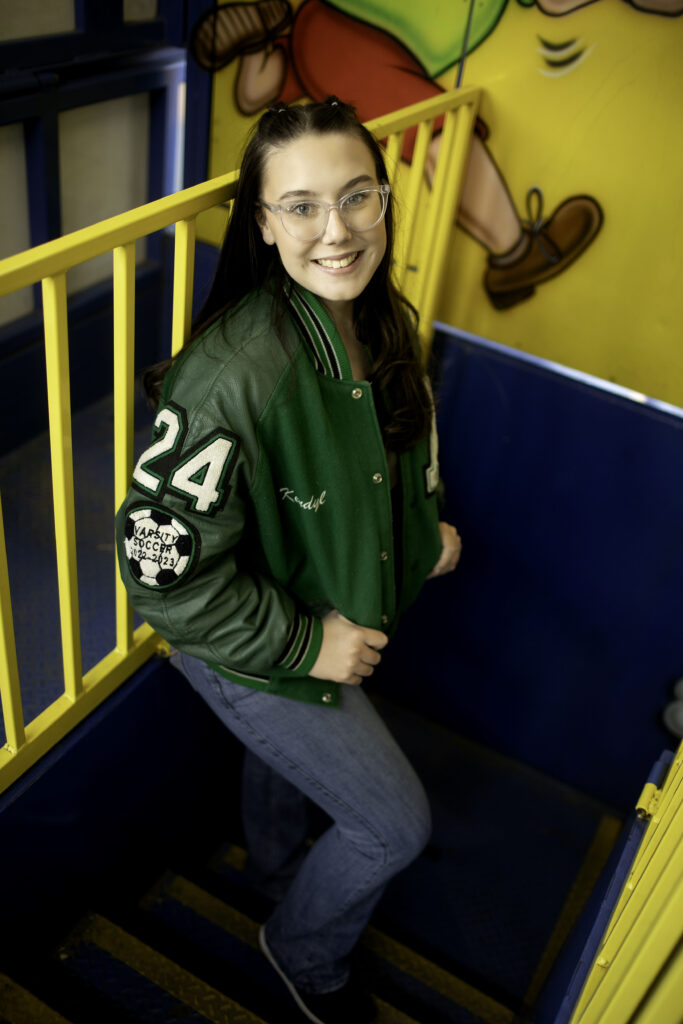 Senior girl posing inside a walk-through funhouse, with playful lighting and bold patterns reflecting around her.