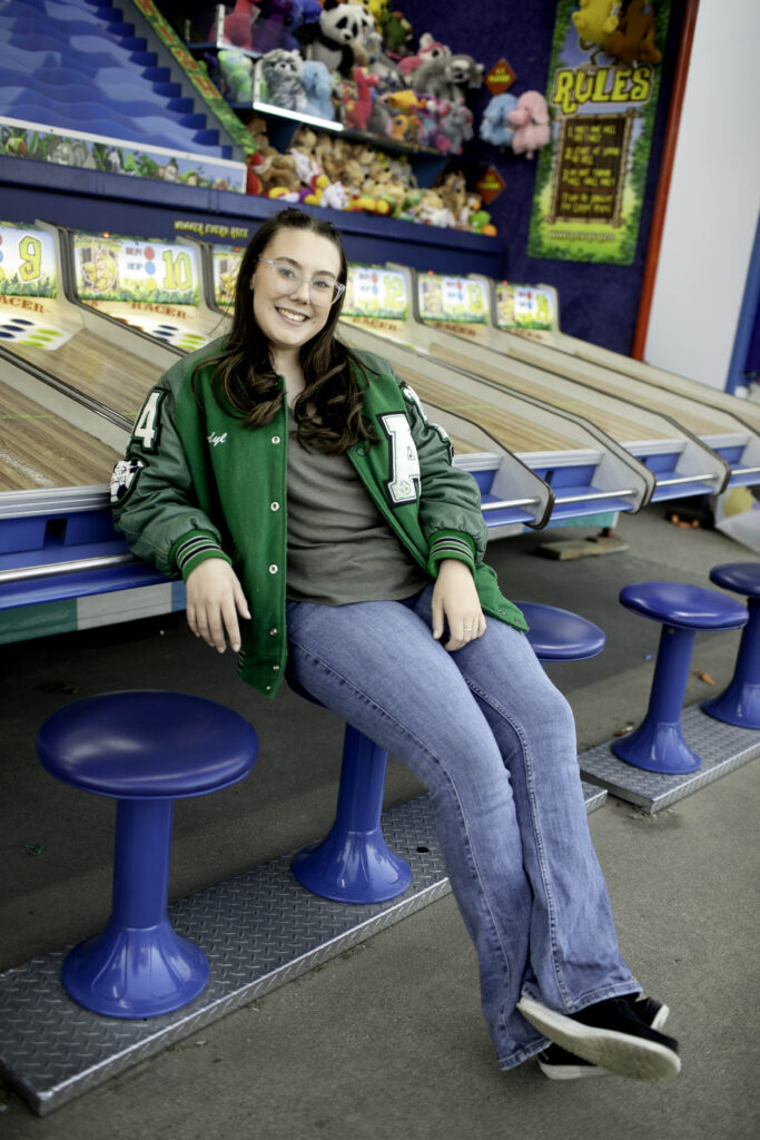 Senior girl leaning on a game counter, smiling as carnival lights reflect in her eyes.