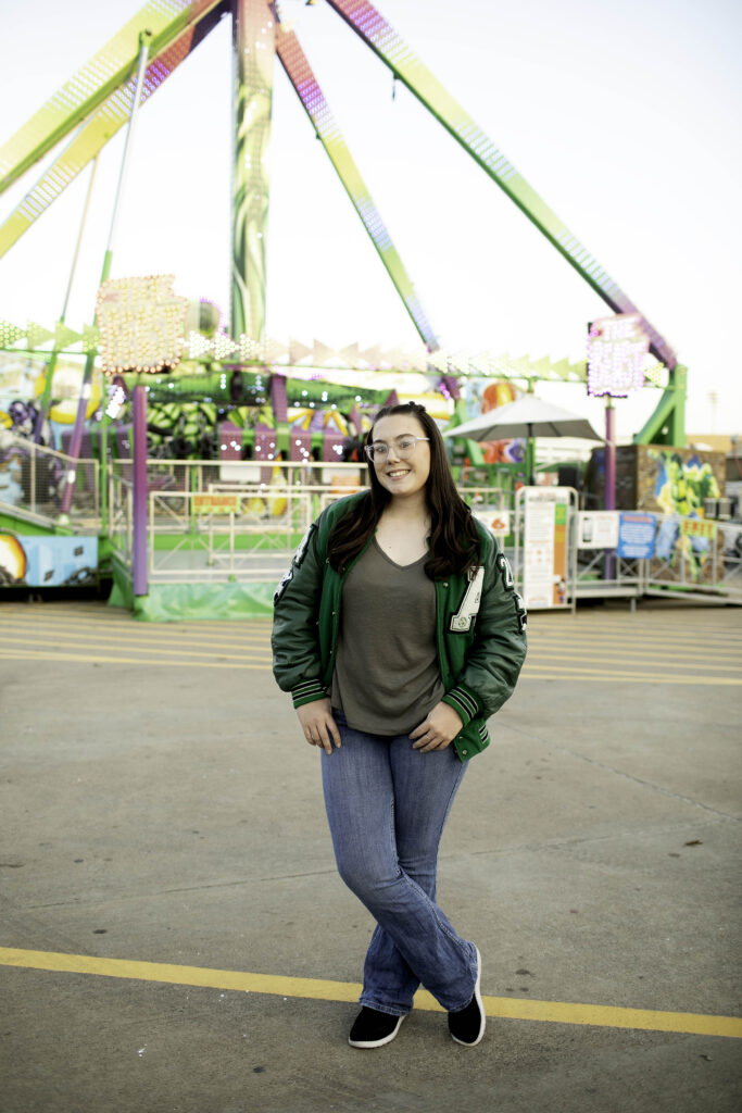 Senior girl standing in front of stuffed animals at a game booth, lights and colors glowing behind her.