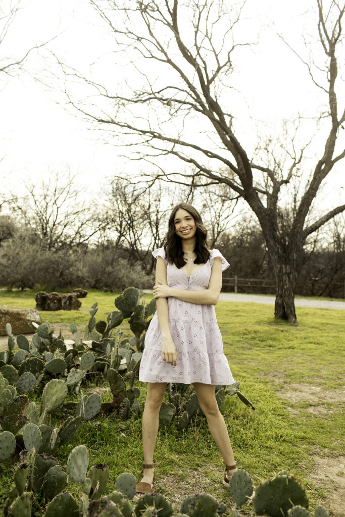 Senior girl with her hands on her hips, posing in front of a large cactus with a confident, poised expression.