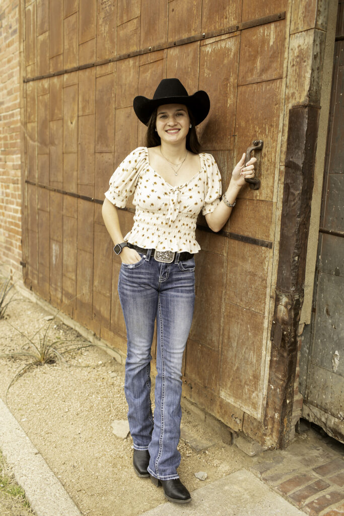 Senior girl laughing with a cowboy hat in hand, walking past the historic buildings of the Fort Worth Stockyards.