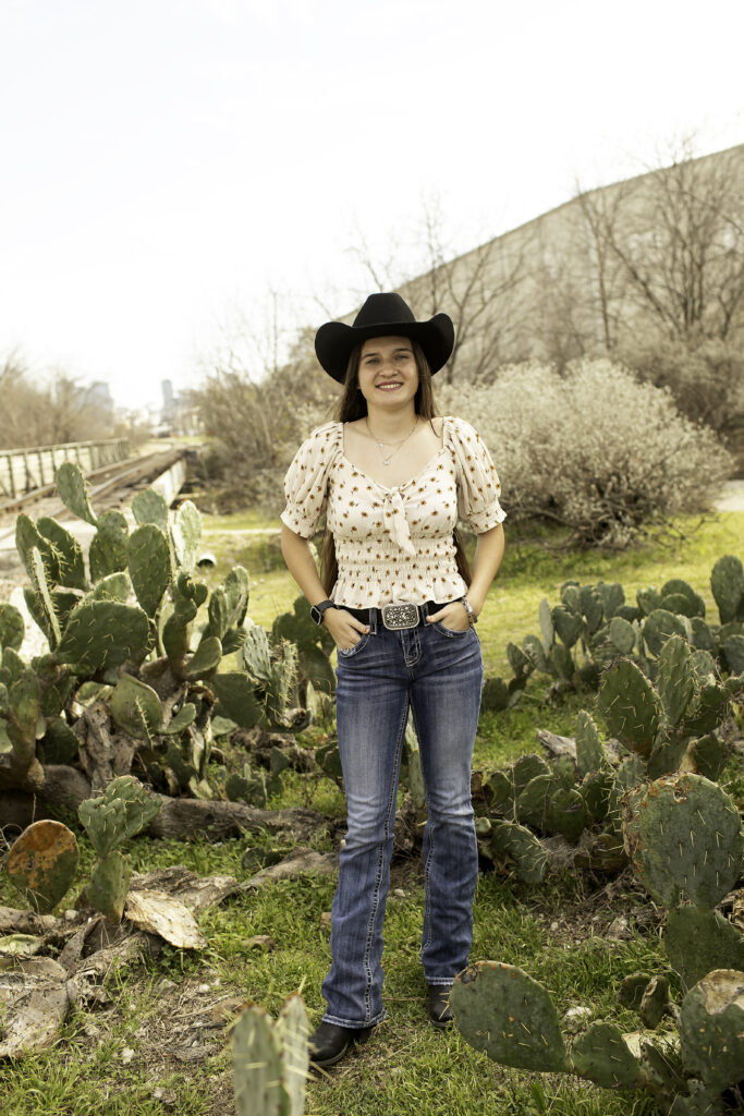 Senior girl with long hair, posing next to a large cactus, dressed in a casual, boho-inspired outfit.