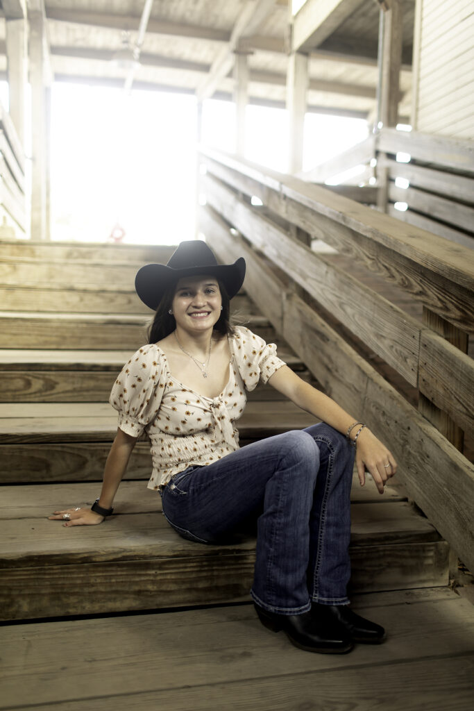 Senior girl holding a cowboy hat, sitting on a wooden rail in the Stockyards with rustic buildings behind her.