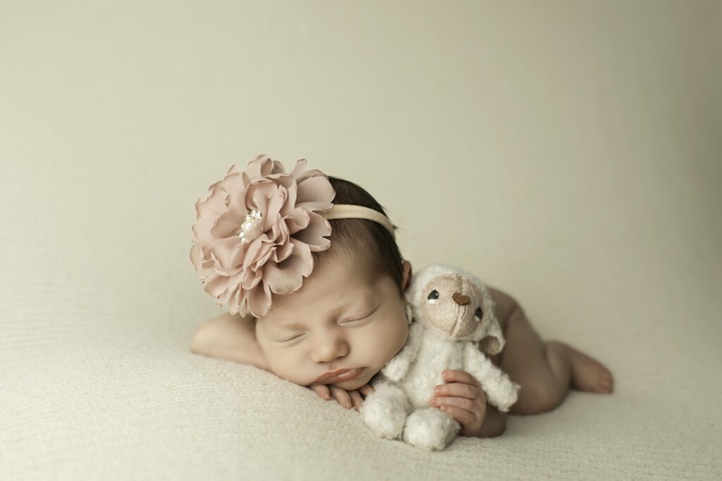 Newborn posed with chin resting gently on her wrist, lying belly-down on a soft white background with a lamb