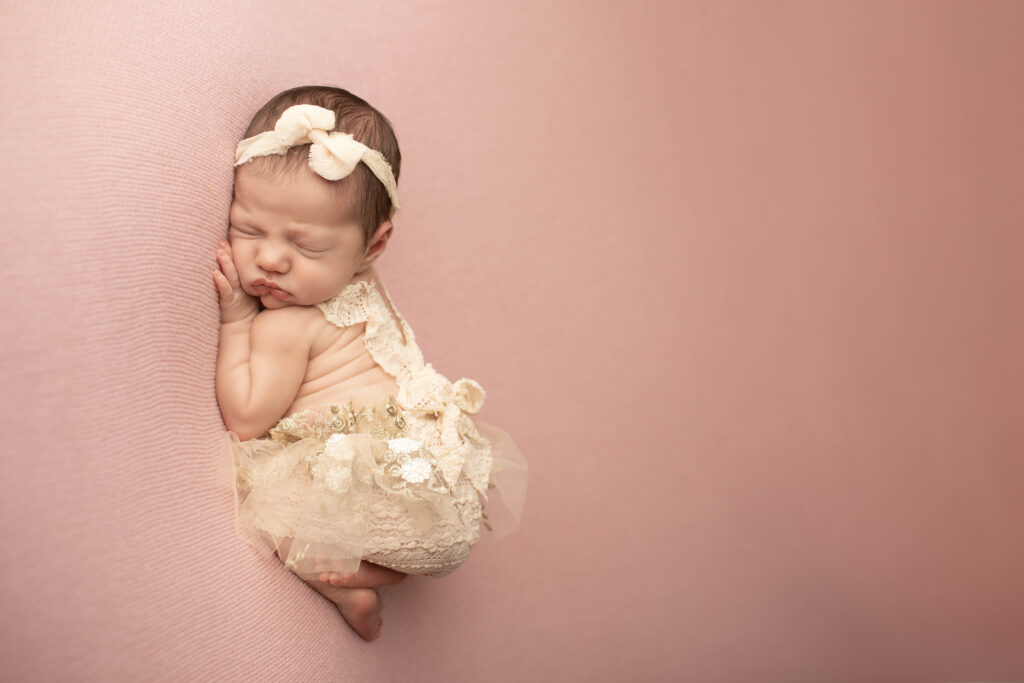 Curled-up newborn girl in the womb pose on a solid pink blanket, arms and legs tucked in close for a cozy look.