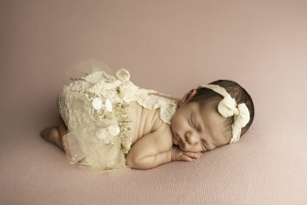 Curled-up newborn girl in the womb pose on a solid pink blanket, arms and legs tucked in close for a cozy look.
