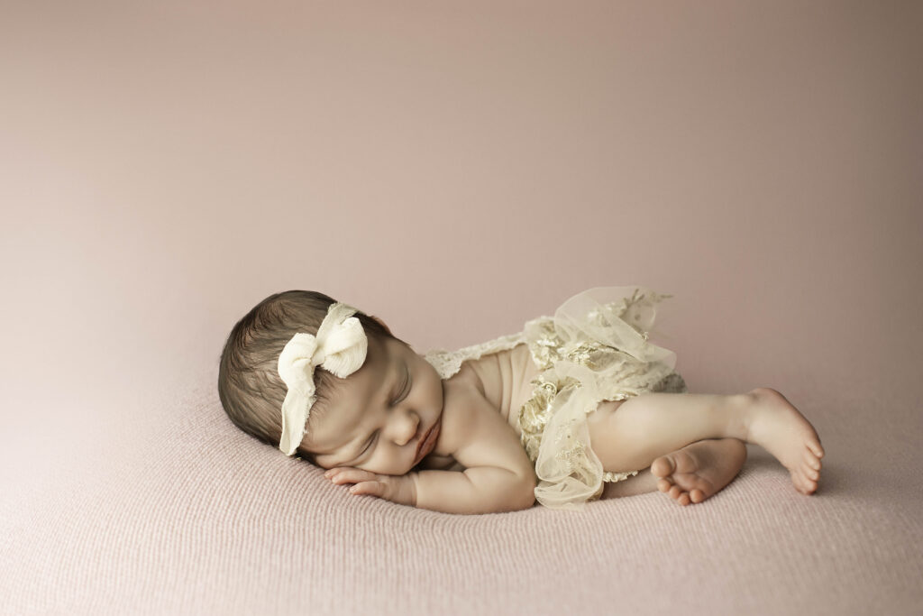 Newborn girl in a side-lying pose on a pink blanket, with hands tucked under her cheek and a soft headband around her head.