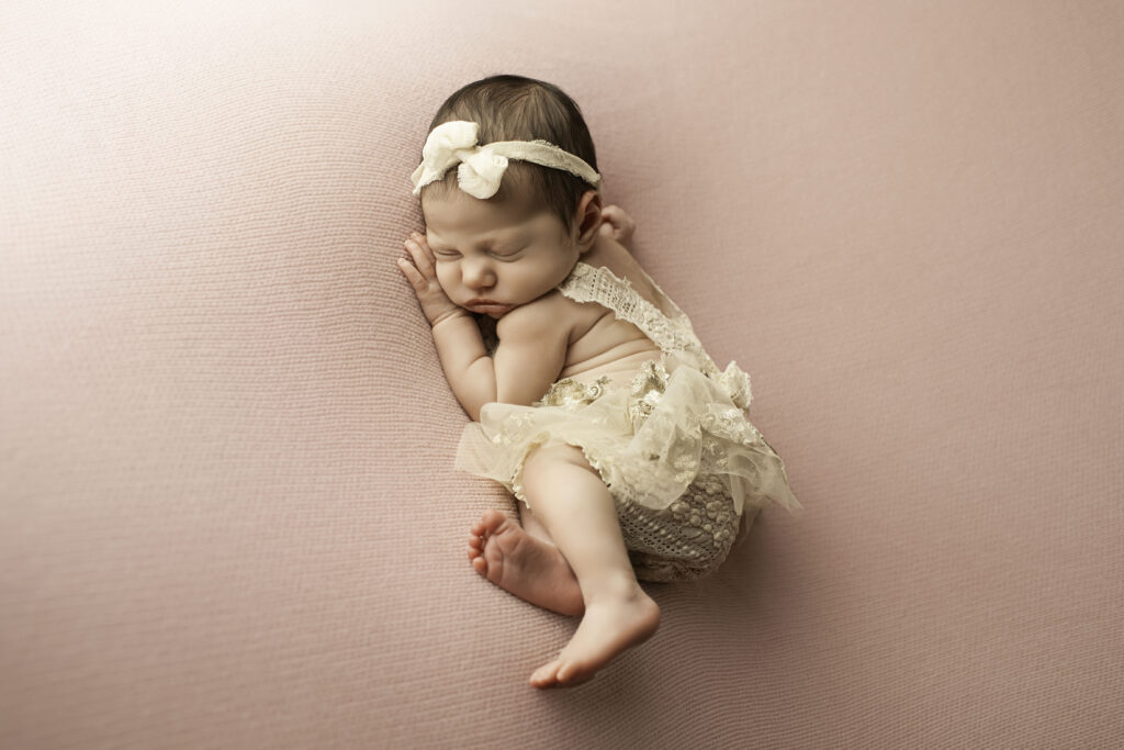 Newborn girl in a side-lying pose on a pink blanket, with hands tucked under her cheek and a soft headband around her head.