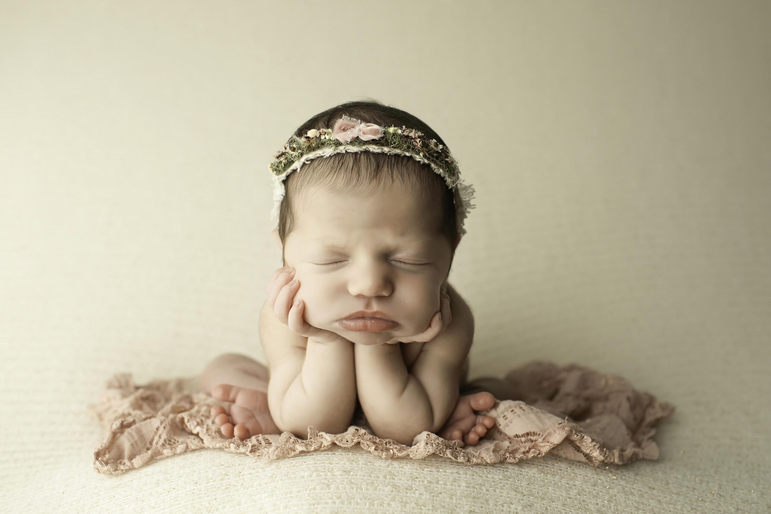 Close-up of a newborn girl in froggy pose on a clean white background, her chin resting in her hands with legs folded beside her.