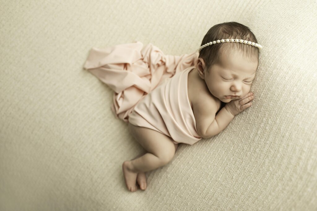 Newborn girl in a side-lying pose on a white blanket, with hands tucked under her cheek and a dainty headband around her head.
