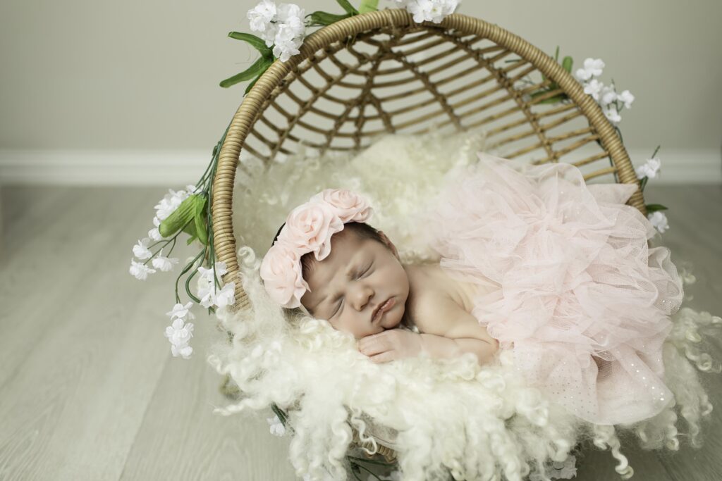 Sleeping newborn girl dressed in a delicate pink tutu, lying in a handcrafted wicker basket with a plush liner, surrounded by soft, textured fabrics in a serene studio environment.
