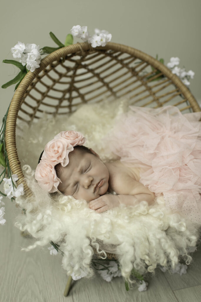 Newborn girl wearing a soft pink tutu, peacefully resting in a woven wicker basket lined with a cozy blanket, set against a neutral-toned studio backdrop.