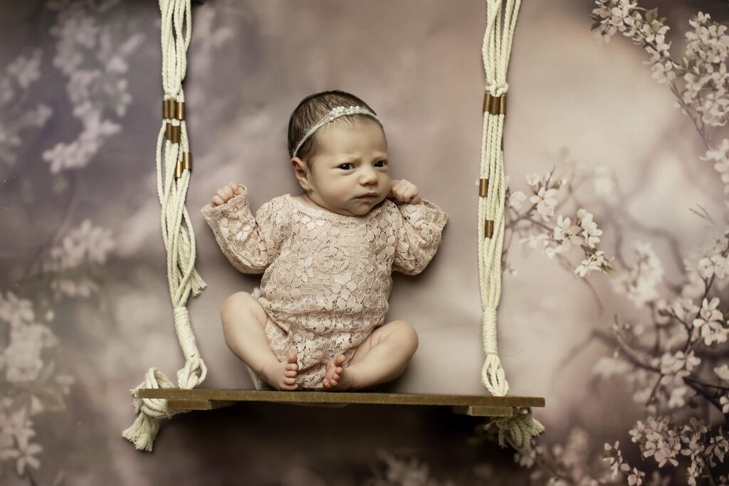 Peaceful newborn girl wrapped in a pale cream lace romper, resting in a handcrafted wooden swing prop with a soft, fringed throw, creating a warm and inviting studio setting.