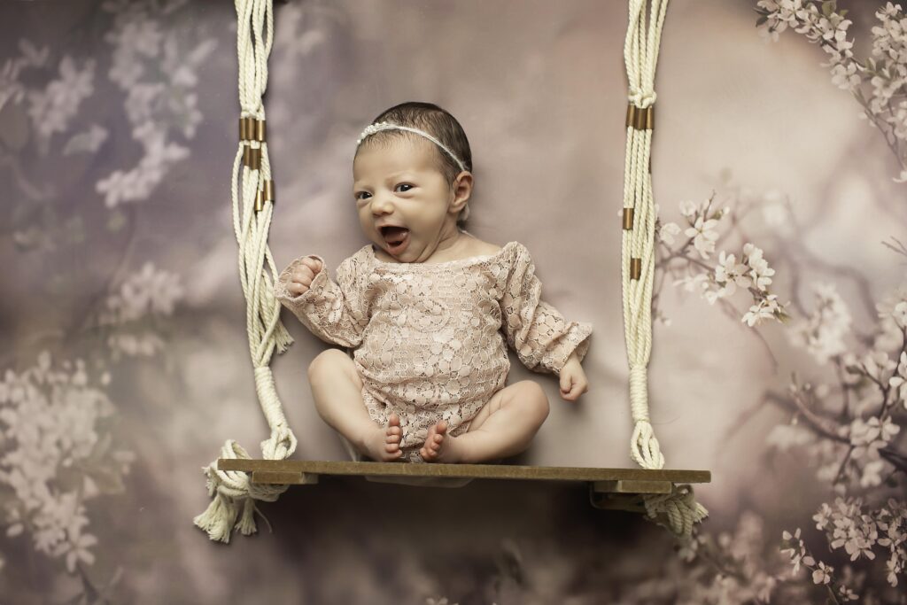 ​Sleeping newborn girl wrapped in a creamy lace romper, lying in a handcrafted wooden swing prop with soft padding, surrounded by soft, textured fabrics in a serene studio environment.
