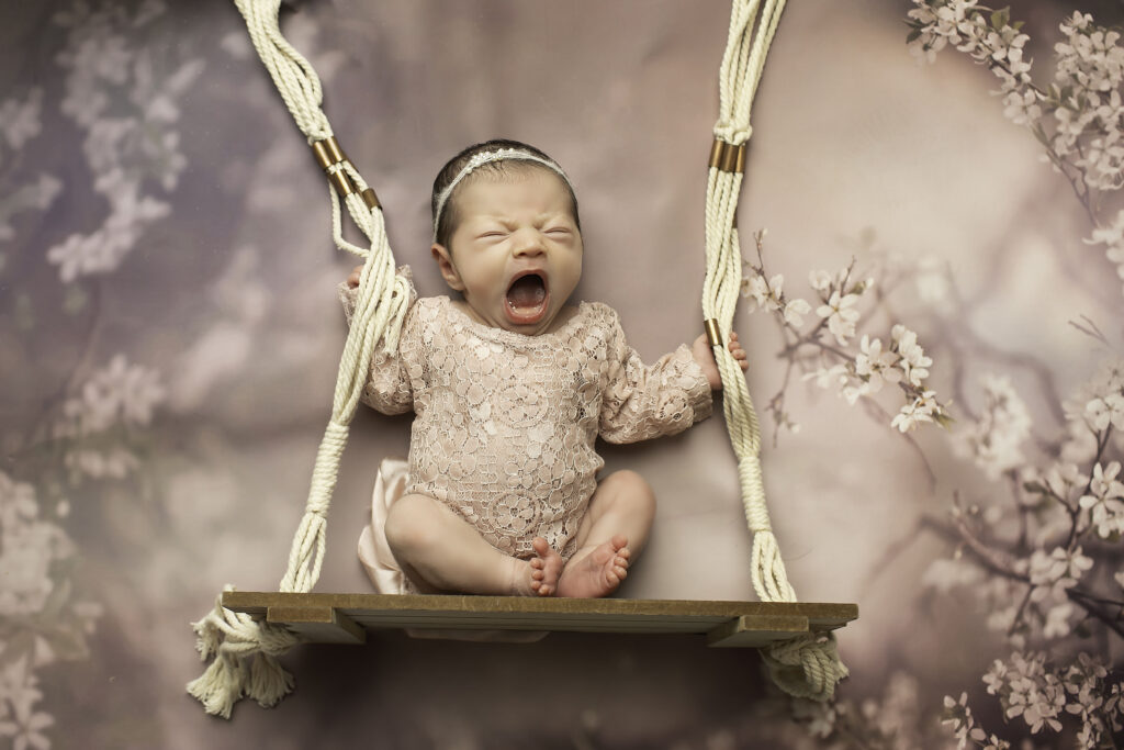 Newborn girl peacefully swaddled in a soft lace romper, resting in a macramé swing prop adorned with delicate tassels, set against a neutral-toned studio backdrop.