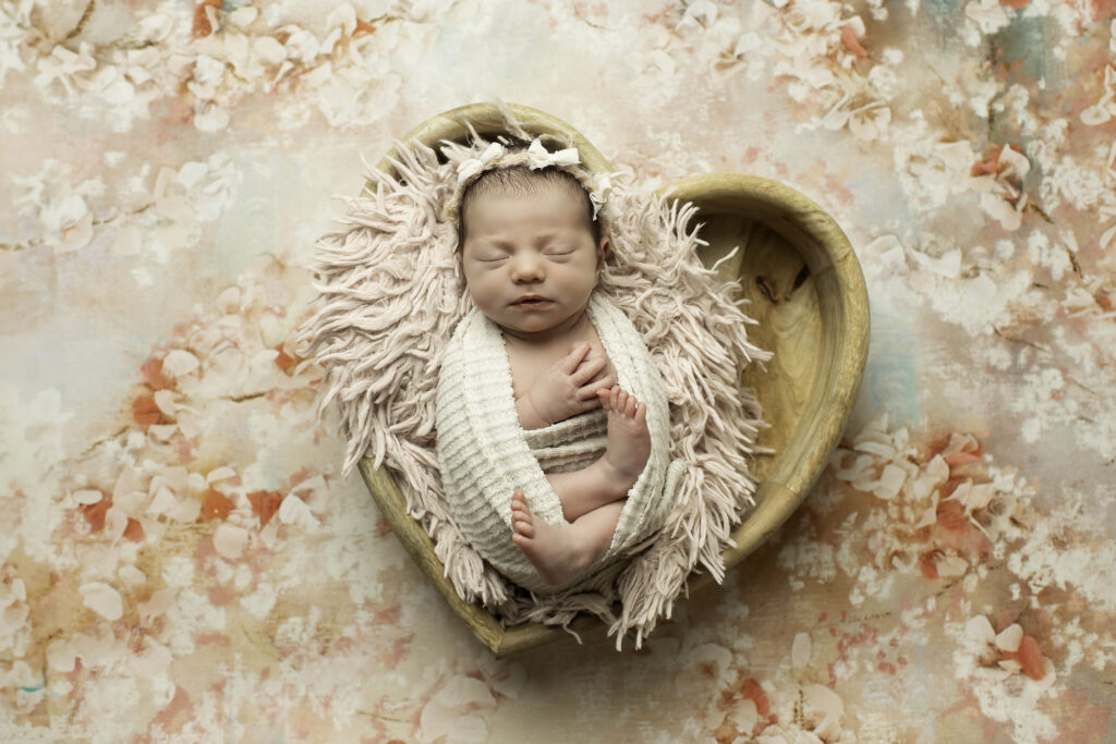 Sleeping newborn girl wrapped in a creamy beige swaddle, lying in a heart-shaped prop lined with a plush faux fur, creating a cozy and serene atmosphere.