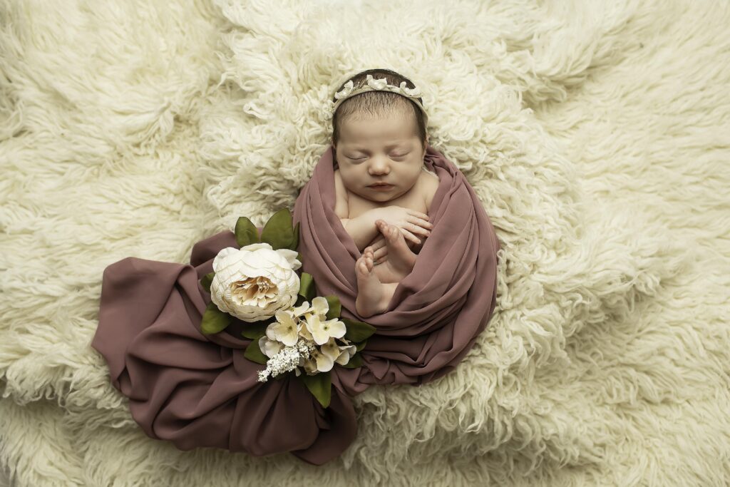 ​Newborn girl swaddled in a soft, dark pink-colored blanket, peacefully sleeping in a cozy studio environment.