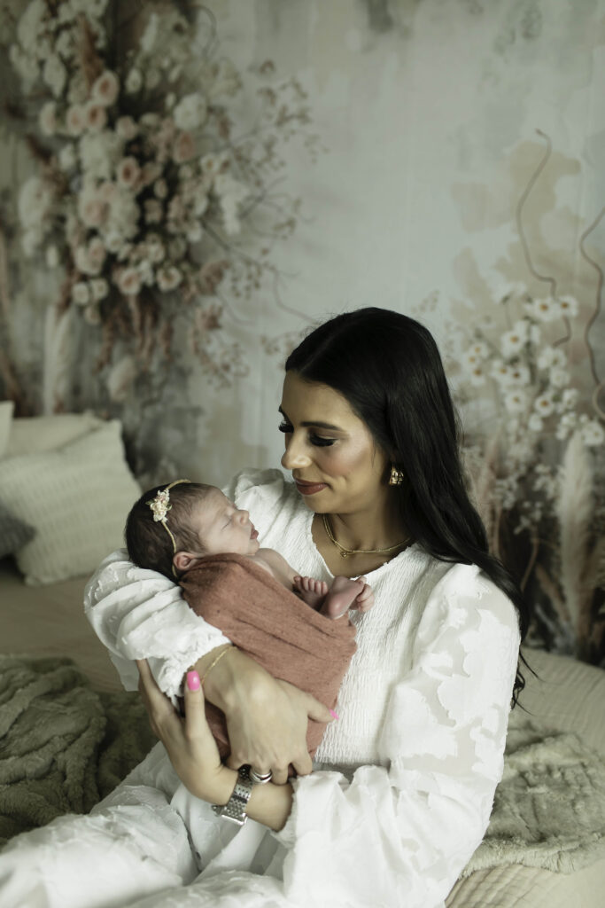 Mom lovingly gazing at her newborn daughter, seated on a boho-style bed adorned with textured pillows and a soft blanket.