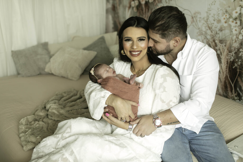 Family of three on a boho-style bed, with the newborn girl swaddled in a soft, earthy-toned blanket, surrounded by a mix of patterned pillows and a macramé wall hanging in the background.