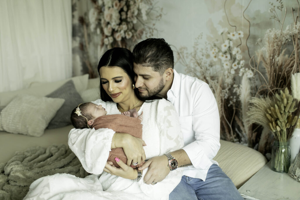 Mom and dad sitting on a bed with a boho-chic design, gently holding their newborn girl, with soft linens and a rattan pendant light casting a warm glow over the scene.