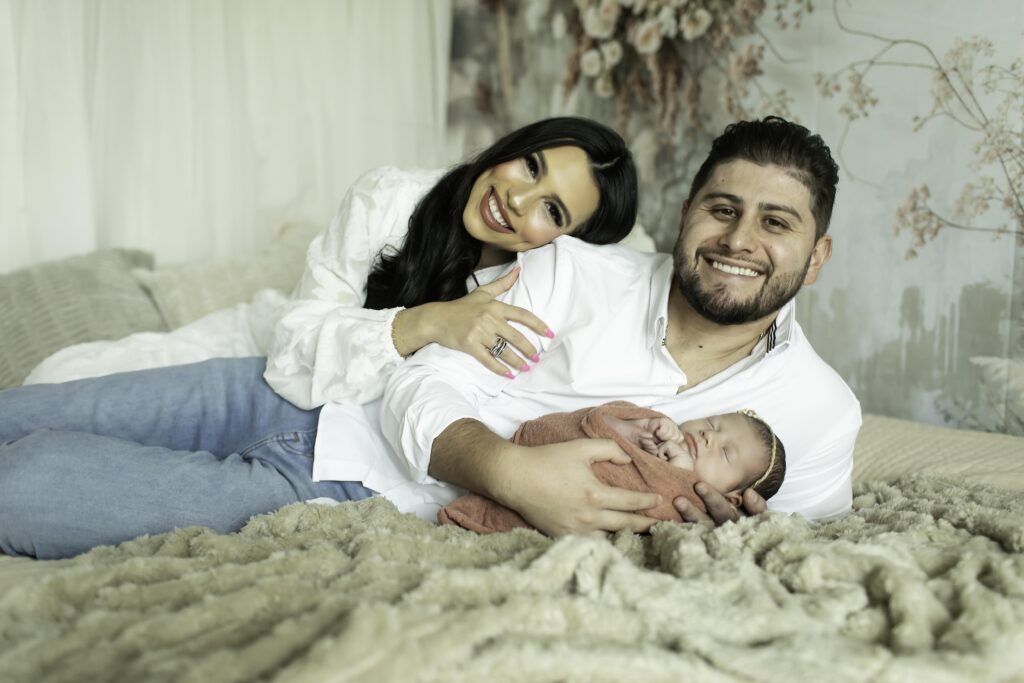 Parents lying on a boho-inspired bed, gently cradling their newborn daughter, with a mix of textured pillows and a soft, neutral-toned blanket creating a cozy atmosphere.