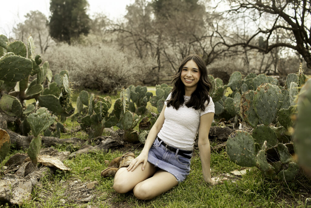 Close-up of senior girl’s face, framed by cactus plants with the warm light of the Stockyards in the background.