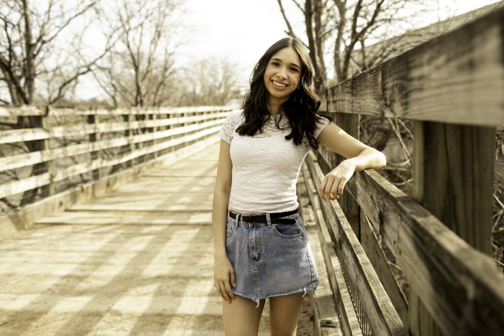 Close-up of senior girl leaning on the wooden railing of the bridge, with the vibrant cacti and desert plants in the Cactus Garden behind her.