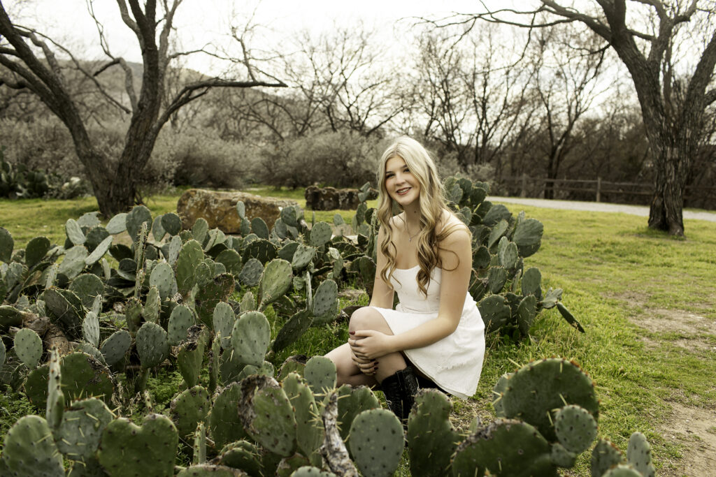 Senior girl in a white dress gazing off into the distance with tall desert plants behind her.