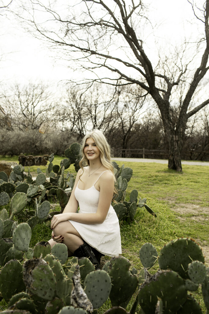Senior girl in white dress smiling softly while framed by large cactus plants and desert flowers.