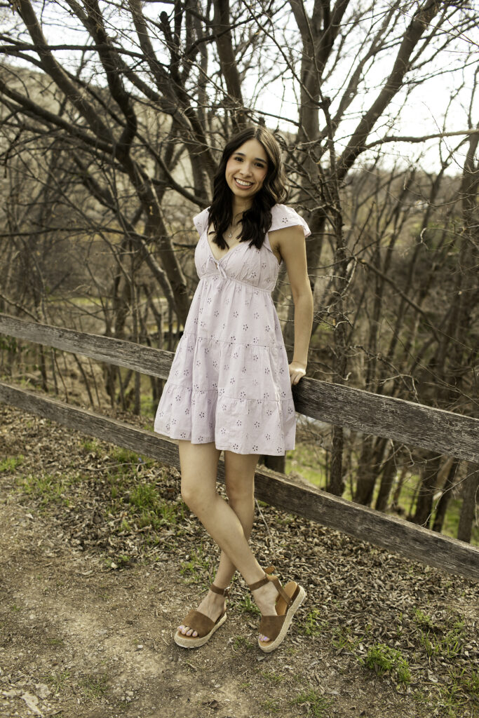 Senior girl perched on a fence rail, looking off into the distance with the warm Texas sun highlighting her face.