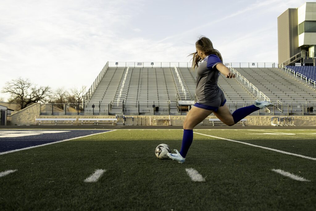 Teen soccer player in uniform executing a powerful kick, with the ball in motion and her body demonstrating dynamic movement on the field.