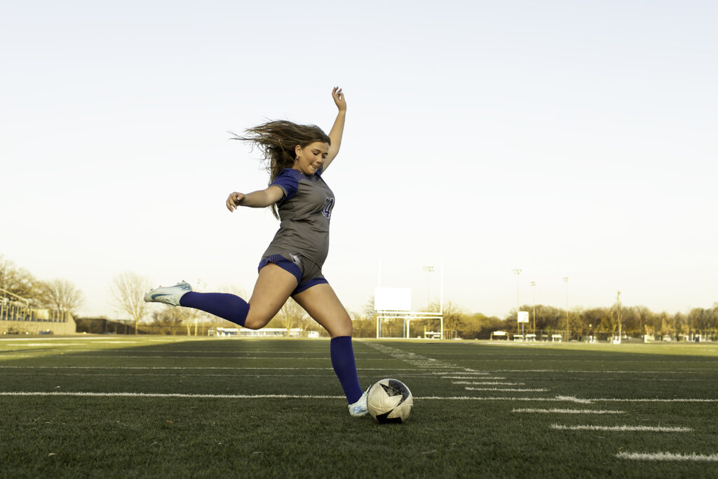 High school soccer player captured mid-kick, her leg extended as she strikes the ball with precision, showcasing athletic form and focus.