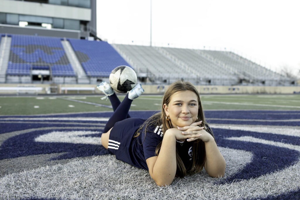 High school soccer player resting a foot on the ball with the bleachers behind her, golden hour lighting glowing.